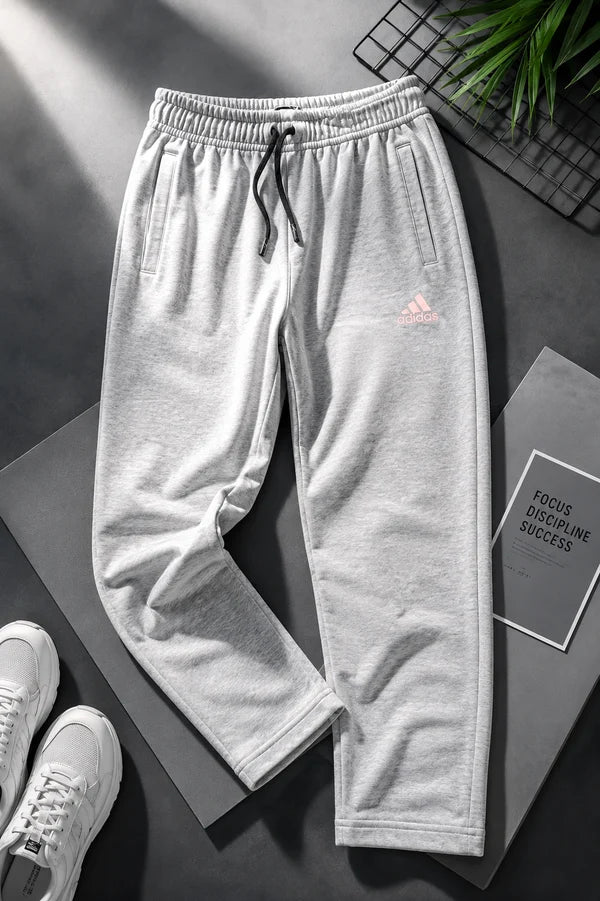 Light gray sweatpants with a drawstring waist and a small pink Adidas logo on the left thigh, styled in a flat-lay with white sneakers, a book, and a plant on a dark gray surface.